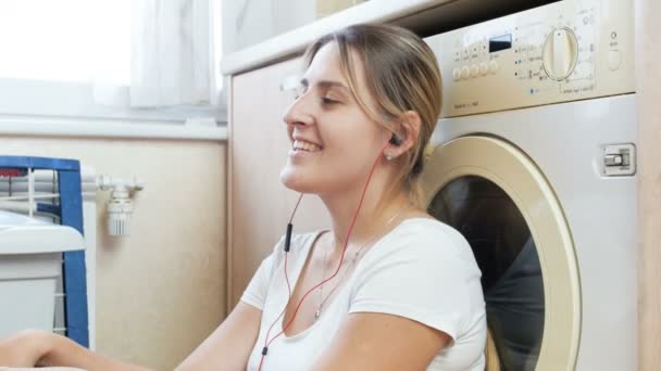 4k filmato di felice giovane donna che ascolta musica in cuffia mentre i vestiti si lavano in lavatrice in lavanderia — Video Stock