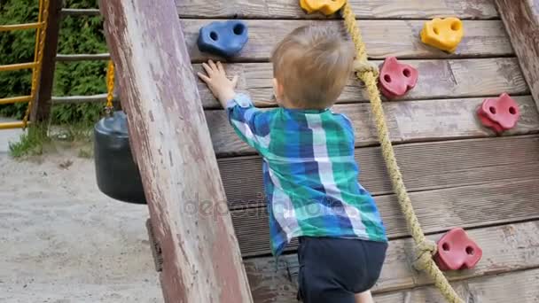 Відео повільного руху малюка, який намагається піднятися на дерев'яну стіну на дитячому майданчику в парку — стокове відео