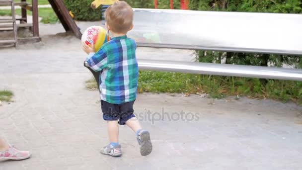 Видео медленного движения маленького мальчика, бегущего и бросающего мяч на детской площадке в парке — стоковое видео