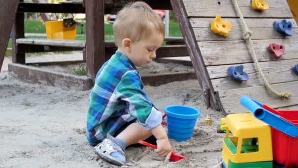 Видео медленного движения симпатичного двухлетнего мальчика, копающего песок на детской площадке в парке — стоковое видео