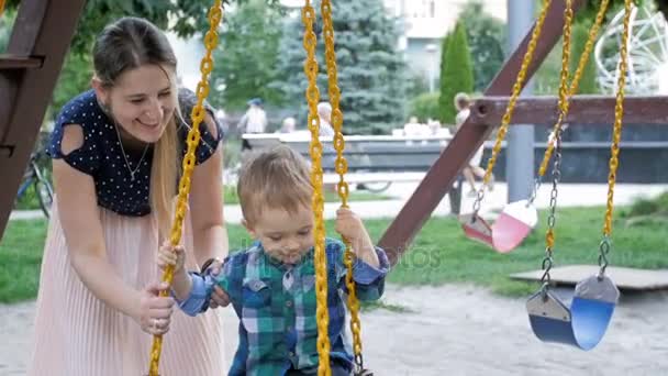 Повільний рух щасливої молодої матері гойдається її маленький син на гойдалці в парку — стокове відео