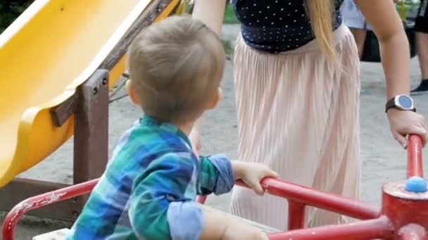 Zeitlupe zeigt junge Mutter, die ihren kleinen Jungen auf buntem Karussell im Park reitet — Stockvideo