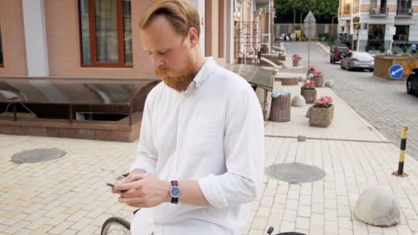 4k imagens de homem barbudo vermelho elegante em camisa branca sentado na bicicleta vintage e digitando mensagem em seu telefone celular — Vídeo de Stock