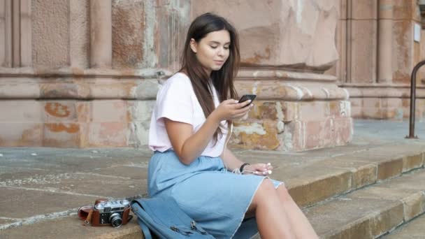 Filmagem 4k de linda garota morena sentada na rua e ouvindo música no smartphone — Vídeo de Stock