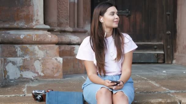 4k-Video einer bezaubernden brünetten Frau, die auf der Straße sitzt und mit Kopfhörern Musik hört — Stockvideo