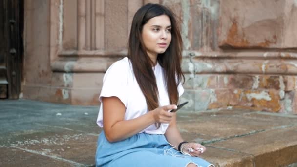 Vídeo em câmera lenta de linda garota morena sentada na rua e inserindo fones de ouvido conectados ao telefone celular — Vídeo de Stock