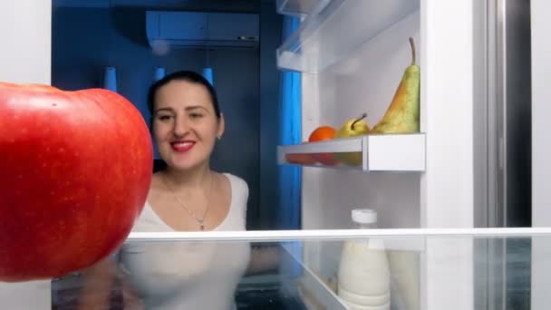 Gros plan vidéo 4k de la jeune femme regardant à l'intérieur du réfrigérateur la nuit et mordant pomme rouge — Video