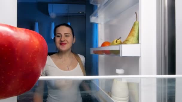 4k кадров, где молодая голодная женщина смотрит в холодильник и ест красное яблоко — стоковое видео