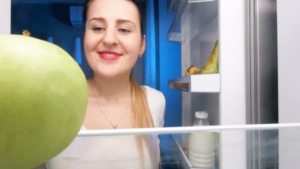 4k video of beautiful smiling woman looking on refrigerator shelves and biting green apple — Stock Video