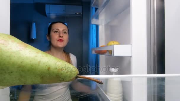4k video av en ung, smilende kvinne som ser inn i kjøleskapet og biter saftig pære – stockvideo