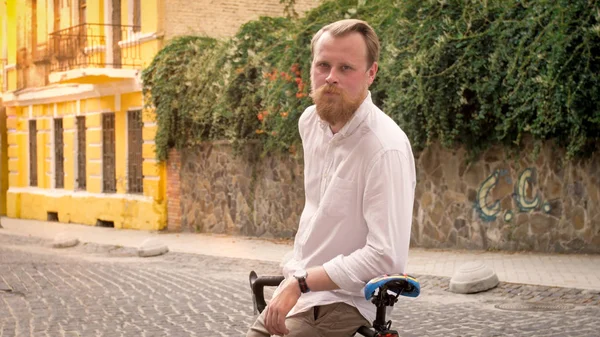 Portrait of stylish bearded man sitting on bicycle on old street at sunset — Stock Photo, Image