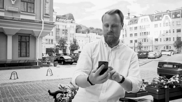 Black and white photo of stylish man with beard leaning on bicycle and using mobile phone