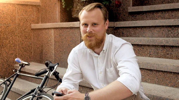 Portrait of smiling stylish man with beard wearing white shirt sitting on stone stairs — Stock Photo, Image