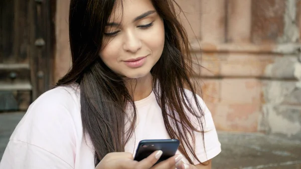 Портрет улыбающейся элегантной девушки с длинными волосами, печатающей сообщения на мобильном телефоне — стоковое фото
