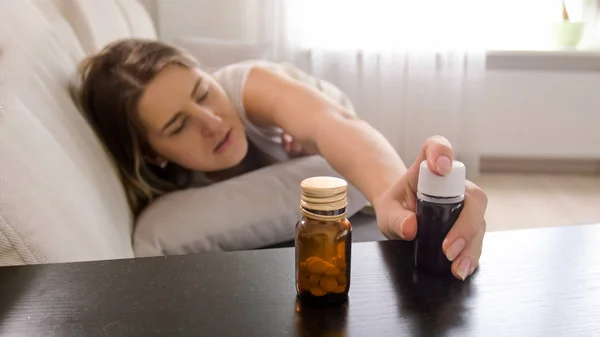 Foto de close-up da jovem mulher doente deitada no sofá e alcançando pílulas na mesa de cabeceira — Fotografia de Stock