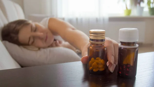 Porträtt av ung sjuk kvinna liggande på sängen och tar flaskan med piller från sängbord — Stockfoto