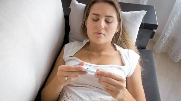 Retrato de mujer joven con gripe tumbada en el sofá y mirando el termómetro —  Fotos de Stock