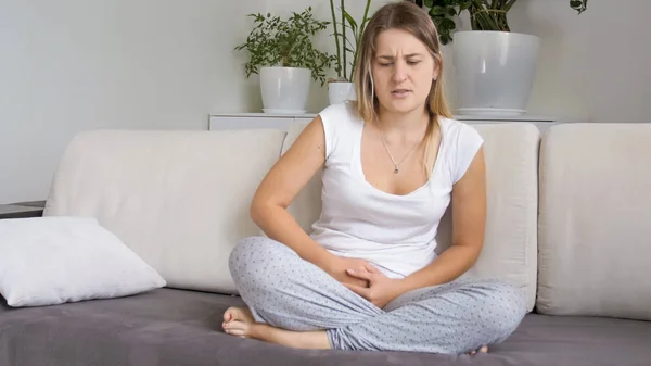 Mujer joven en pijama sentada en el sofá y sufriendo de dolor menstrual —  Fotos de Stock