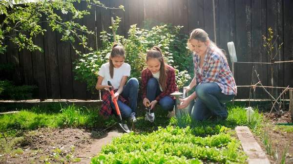 Foto de madre joven con hijas sentadas en el jardín y cuidando de la cama del jardín — Foto de Stock