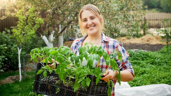 Tonas foto av unga leende kvinna som bär låda med plantor i trädgården — Stockfoto