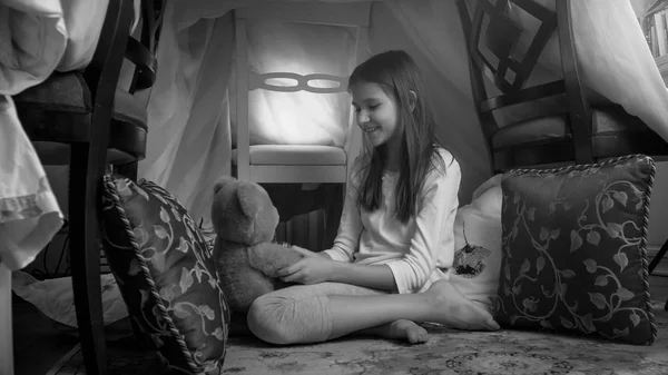 Черно-белое фото милой девушки, играющей с плюшевым мишкой в палатке-вигваме дома — стоковое фото