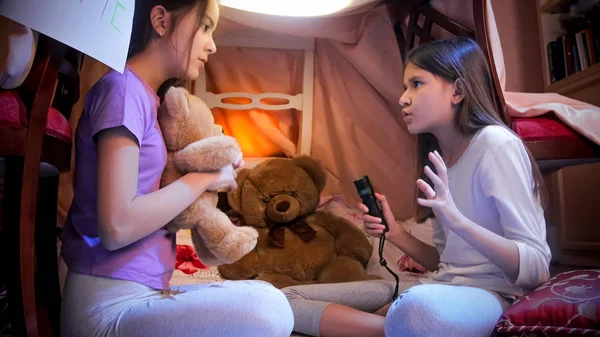 Linda chica en pijama contando historia de miedo a su amigo en tienda de campaña hecha a sí mismo en el dormitorio —  Fotos de Stock