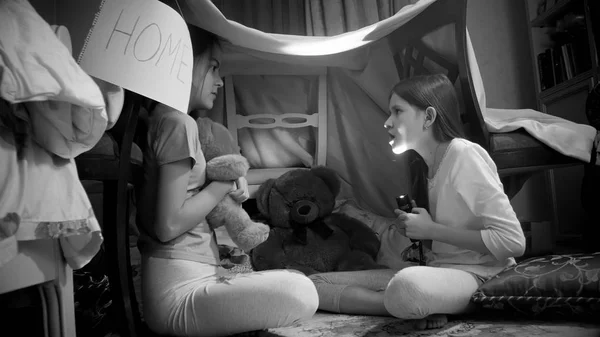 Black and white photo of two girls telling scary stories with flashlight in tent at bedroom — Stock Photo, Image