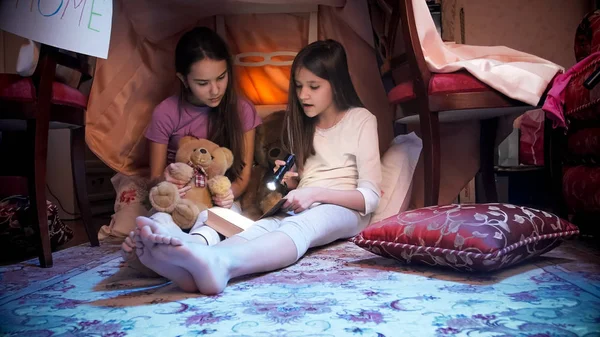 Two sisters in pajamas sitting in tepee tent in bedrom at night and reading book — Stock Photo, Image