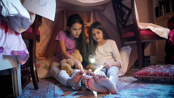 Two girls in pajamas sitting on floor at bedroom and reading book with flashlight — Stock Photo, Image