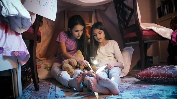 Two girls in pajamas sitting in tepee tent at bedroom and reading book with flashlight — Stock Photo, Image