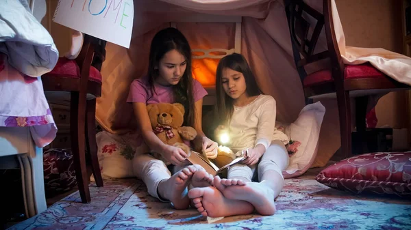 Two girls wearing pajamas with flashlight and book in selfmade tent at bedroom — Stock Photo, Image