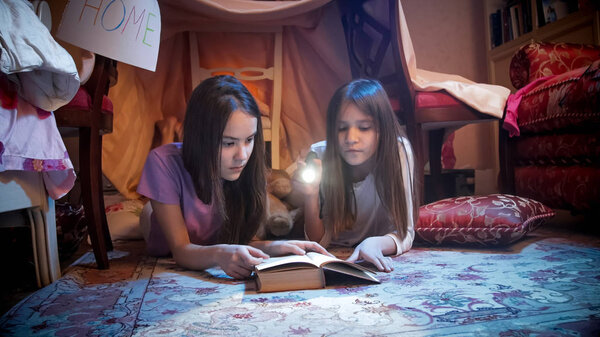 Portrait of two girls in pajamas lying on floor at bedroom and reading book at night