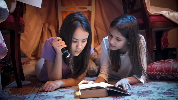 Portrait of two cute sisters lying in tepee tent at bedroom and reading book with flashlight — Stock Photo, Image