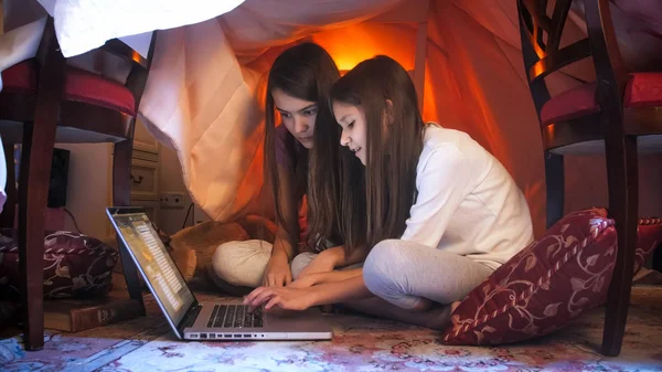 Två söta tjejer i pyjamas som sitter med laptop i tält gjort av filtar — Stockfoto