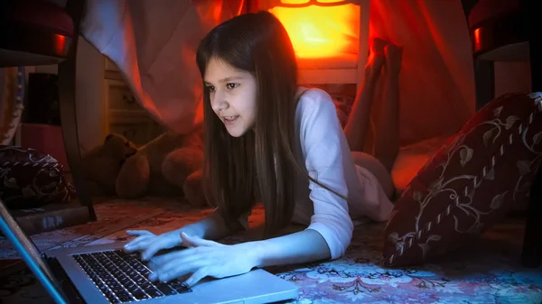 Linda chica sonriente acostada en el suelo por la noche y utilizando el ordenador portátil — Foto de Stock
