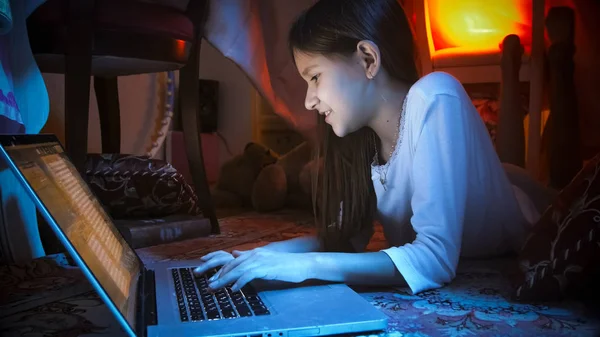 Retrato de menina sorridente em pijama deitado no chão e digitando mensagem no laptop — Fotografia de Stock