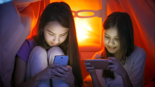 Pijama smartphones battaniyenin altında geceleri kullanarak iki kız kardeş portresi — Stok fotoğraf