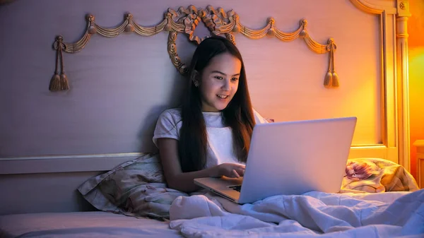 Retrato de una chica sonriente acostada en la cama y usando el portátil por la noche —  Fotos de Stock