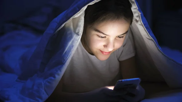 Porträtt av tonårsflicka att skriva meddelande på smartphone under filt på natten — Stockfoto