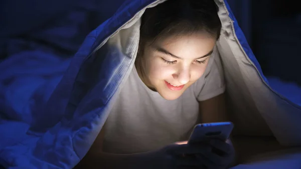 Ritratto di ragazza adolescente sorridente messaggistica sul telefono cellulare sotto coperta di notte — Foto Stock