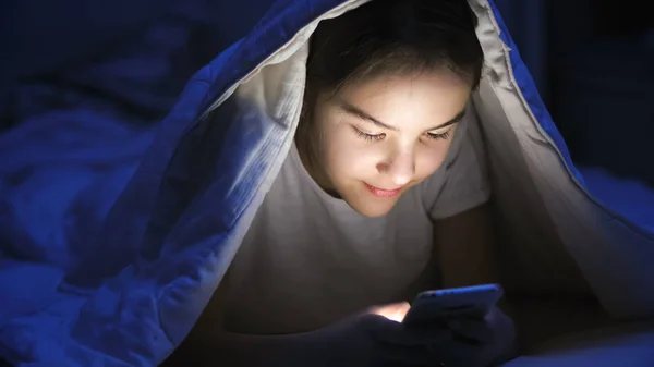 Porträtt av tonårig flicka liggande under filt med mobiltelefon — Stockfoto