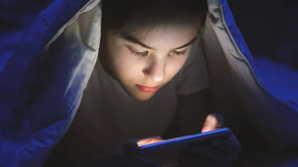 Detailní portrét dívky, ležící pod deku a hraní na mobilu — Stock fotografie