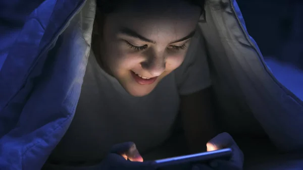 Porträtt av leende flicka surfa sociala media på mobiltelefon under filt på natten — Stockfoto