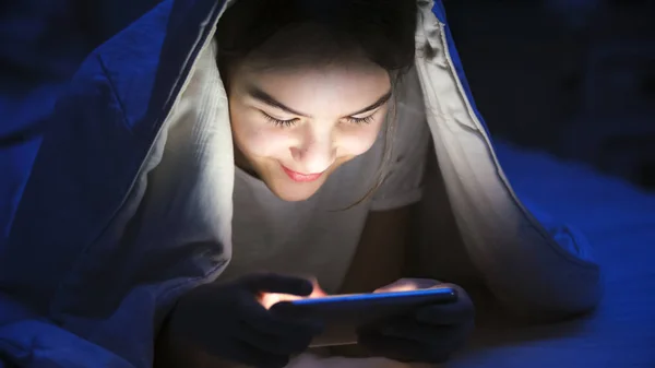 Porträtt av leende vacker flicka att skriva meddelande på smartphone under filt på natten — Stockfoto