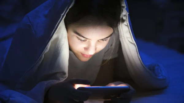 Geceleri battaniye altında hareket eden telefon ile gülümseyen kız closeup portresi — Stok fotoğraf