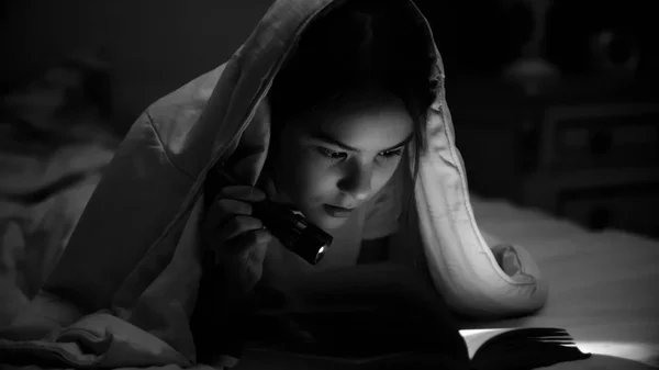 Black and white photo of girl lying under blanket and reading book with flashlight — Stock Photo, Image