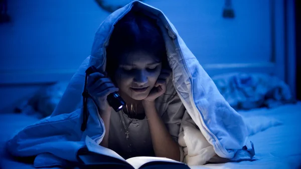 Sonriente niña leyendo libro en dormitorio oscuro antes de ir a dormir —  Fotos de Stock