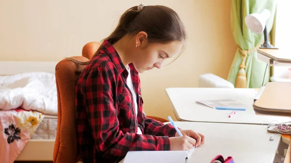 Retrato de menina bonita escrevendo em caderno atrás da mesa no quarto — Fotografia de Stock