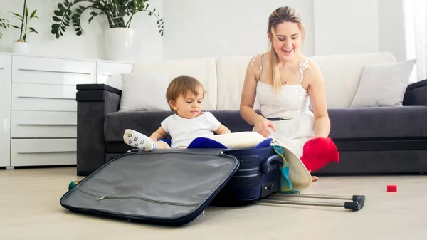 Cute toddler boy sitting in suitcase while mother packing things in suitcase for holiday — Stock Photo, Image