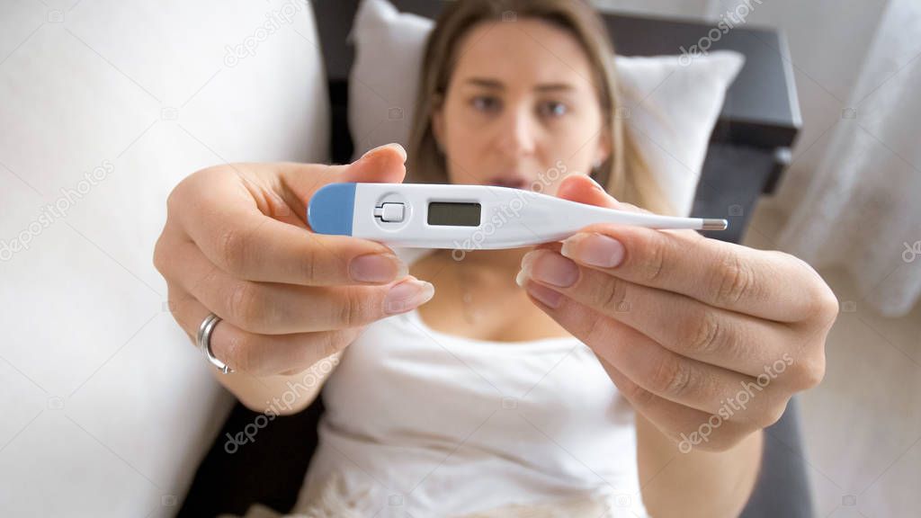 Closeup photo of young sick woman lying on sofa and showing thermometer in camera